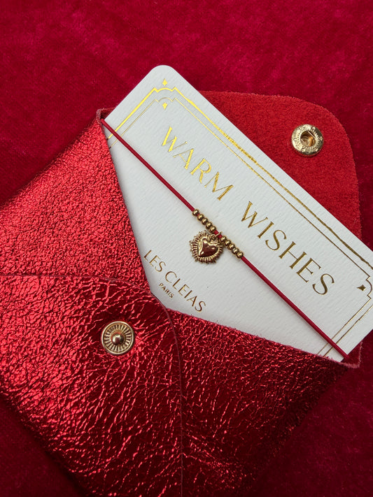 Gold bracelet on a 'Warm Wishes' card with 'LES CLEIAS' branding on a red textured surface.