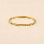 A gold-plated bangle bracelet with a row of small green and light blue enamel dots, displayed on a textured beige background.