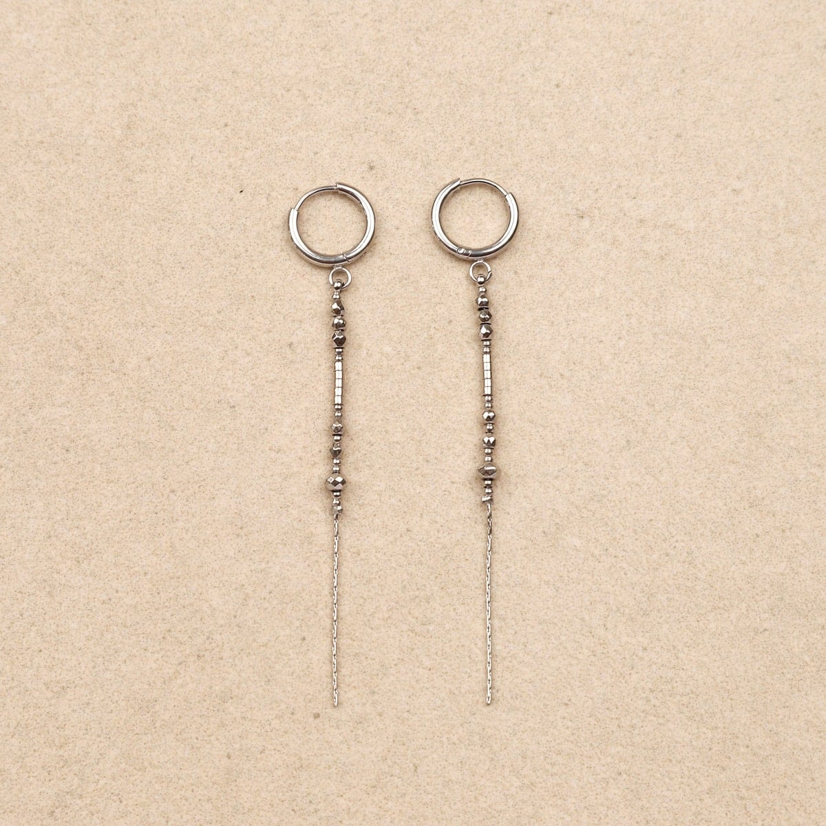 A pair of silver-toned dangle earrings with circular hoop tops, small bead details, and thin chains, displayed on a textured beige background.