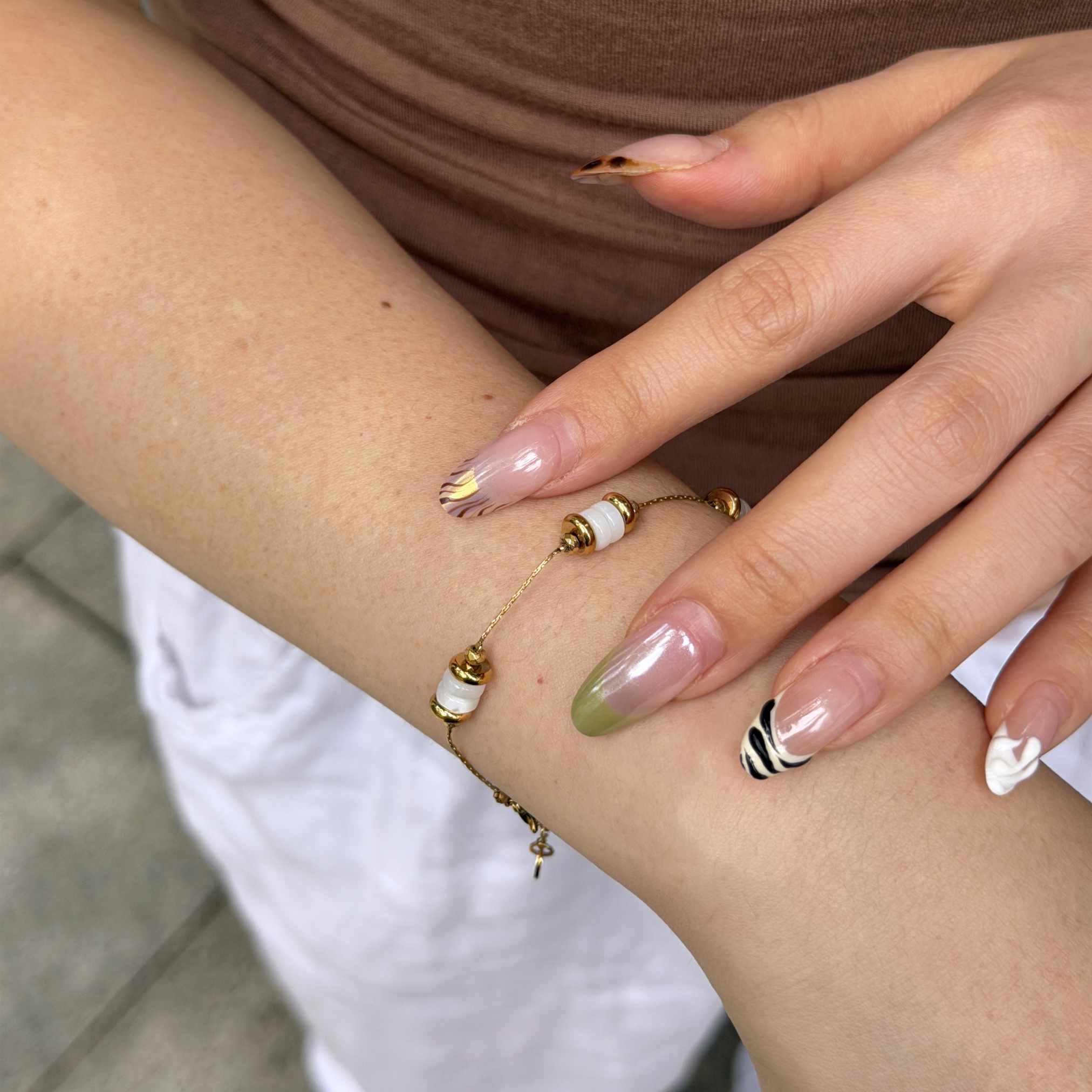 A close-up of a person's wrist and hand wearing a delicate gold chain bracelet with white and gold beads, styled with manicured nails.
