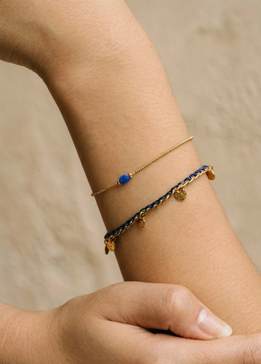 Close-up of a wrist wearing two gold bracelets with blue accents on a neutral background