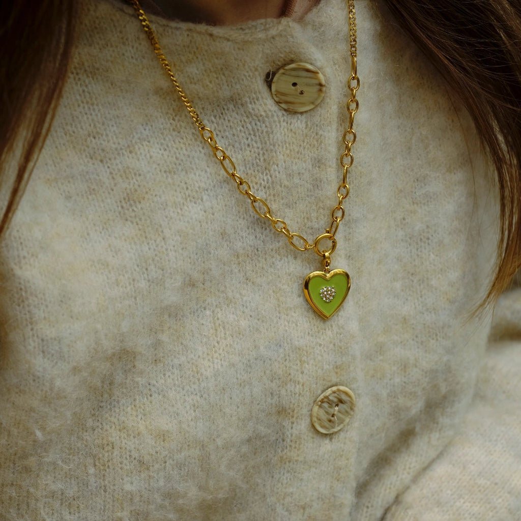 Close-up of a person wearing a gold necklace with a green heart pendant on a beige textured background.