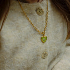 Close-up of a person wearing a gold necklace with a green heart pendant on a beige textured background.