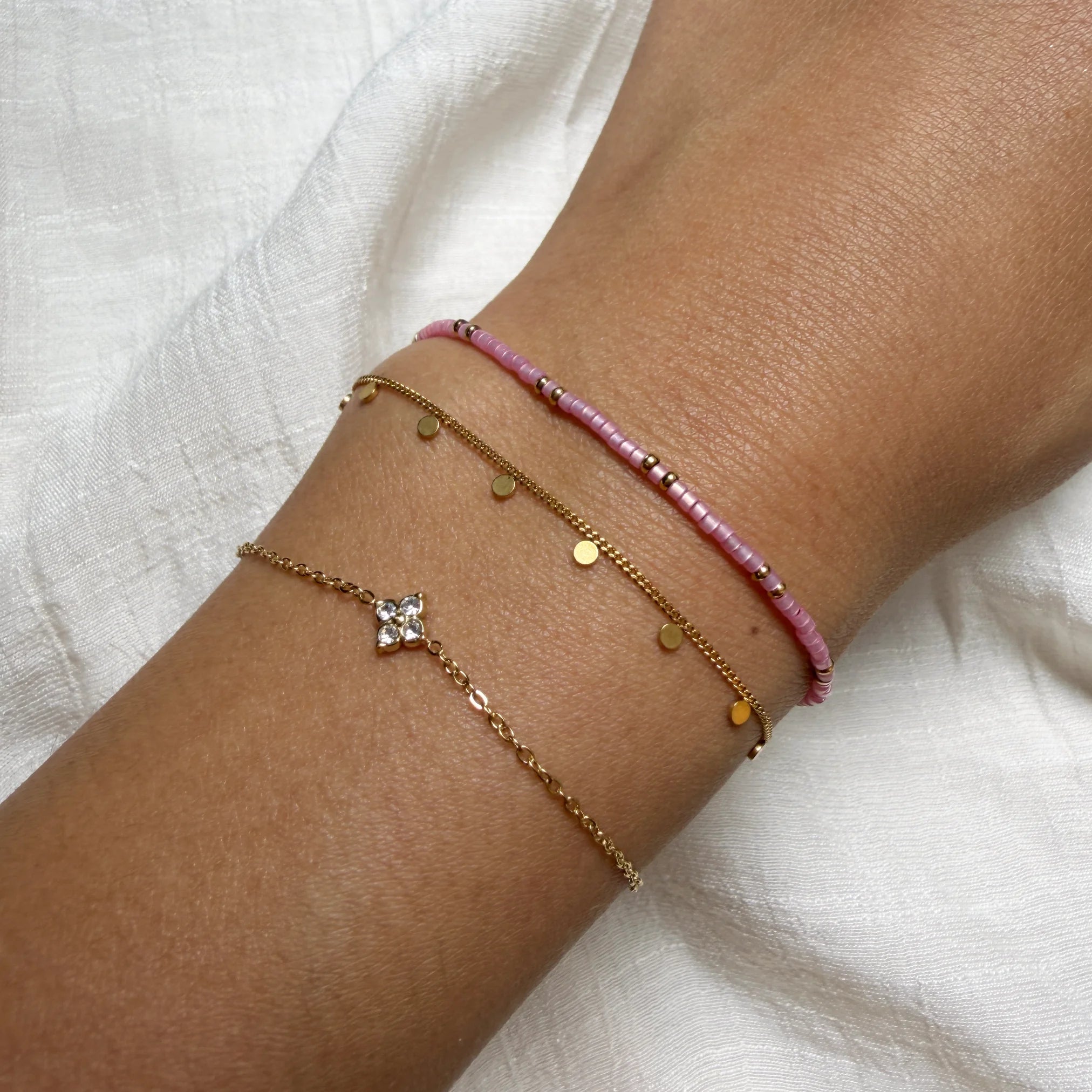 Close-up of a wrist wearing multiple gold and pink beaded bracelets on a white fabric background.