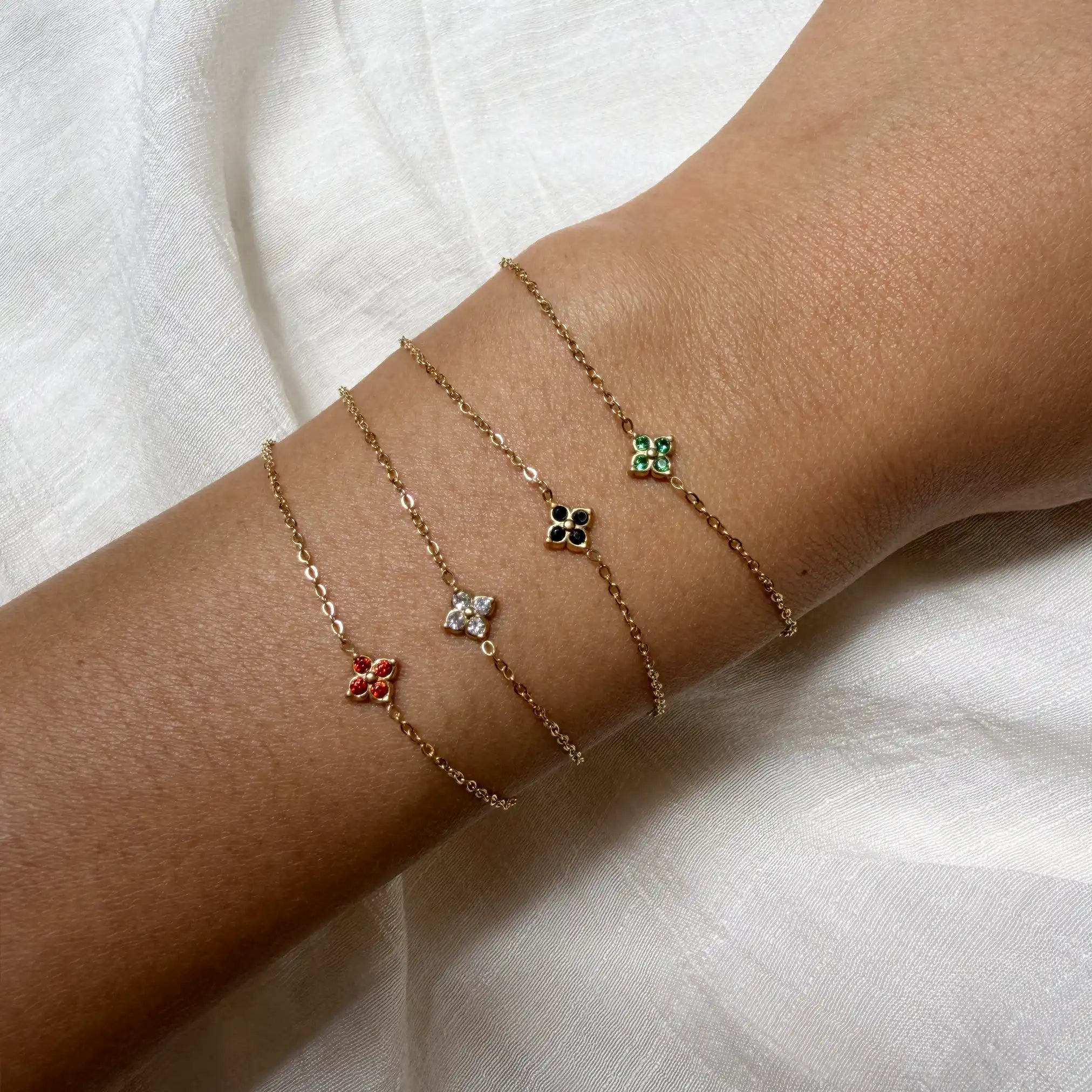 Three delicate gold anklets with floral charms on a white fabric background