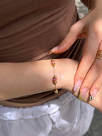 A close-up of a delicate gold chain bracelet with pink and orange bead accents worn on a woman's wrist, styled with a brown top and white skirt.