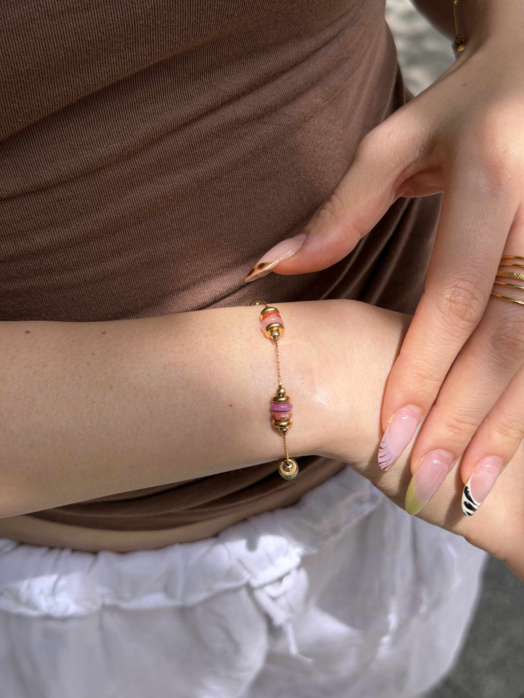 A close-up of a delicate gold chain bracelet with pink and orange bead accents worn on a woman's wrist, styled with a brown top and white skirt.