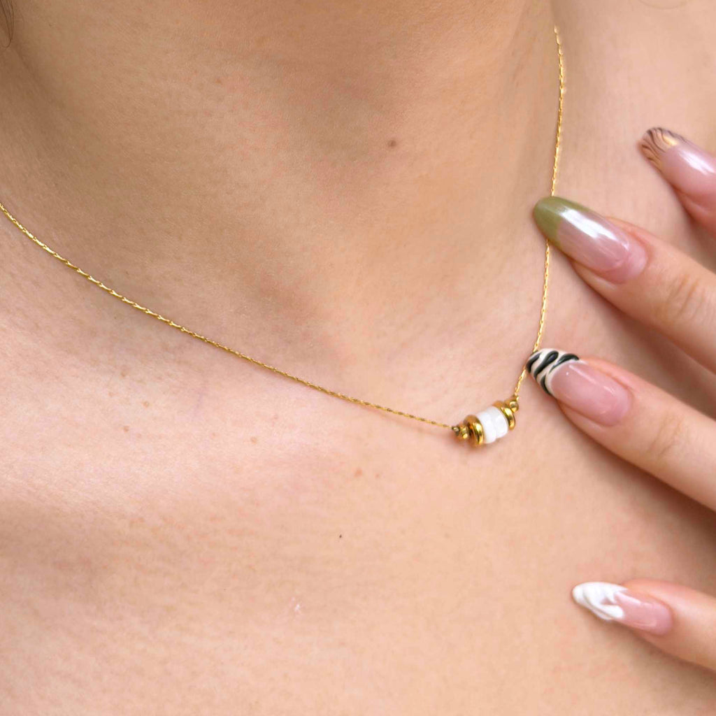 A close-up of a delicate gold chain necklace worn on a model's neck, featuring a small gold bead, a white bead, and a black-and-white striped bead accent.
