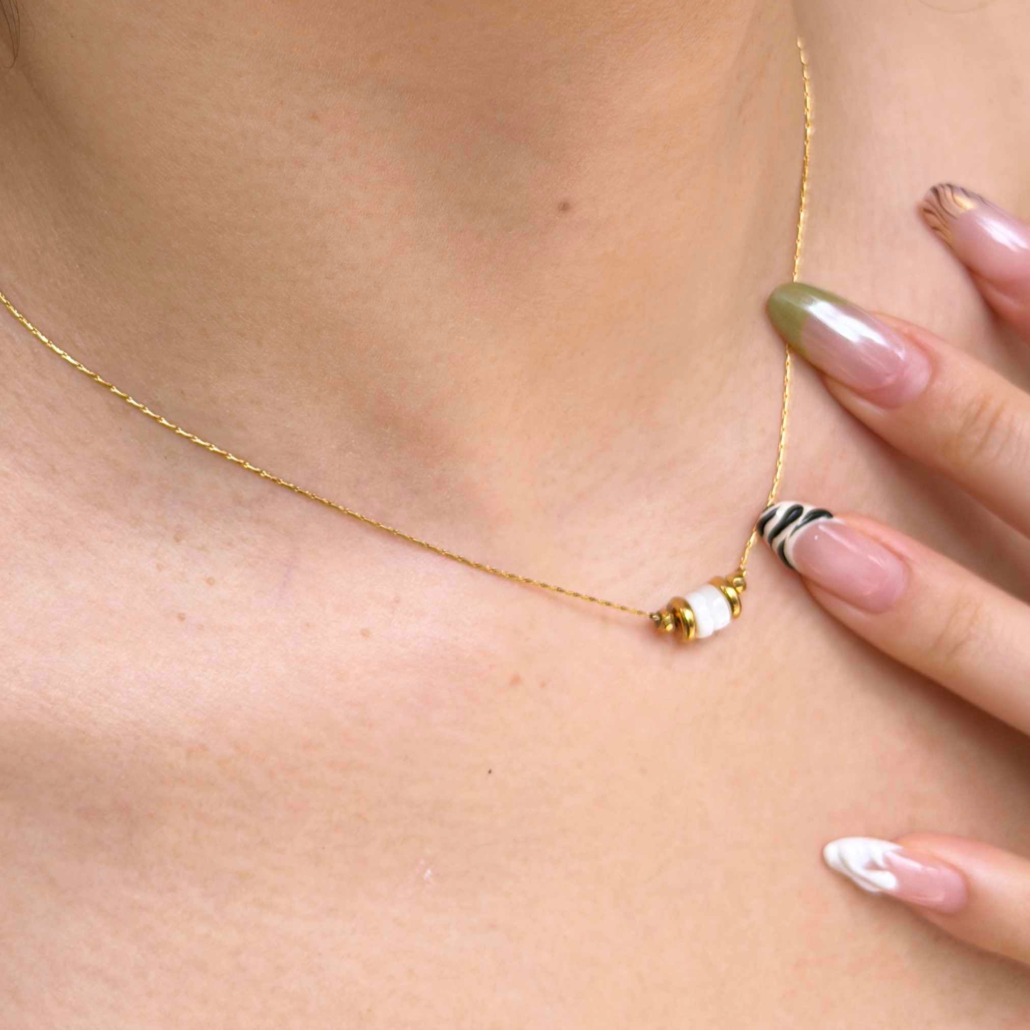 A close-up of a delicate gold chain necklace worn on a model's neck, featuring a small gold bead, a white bead, and a black-and-white striped bead accent.