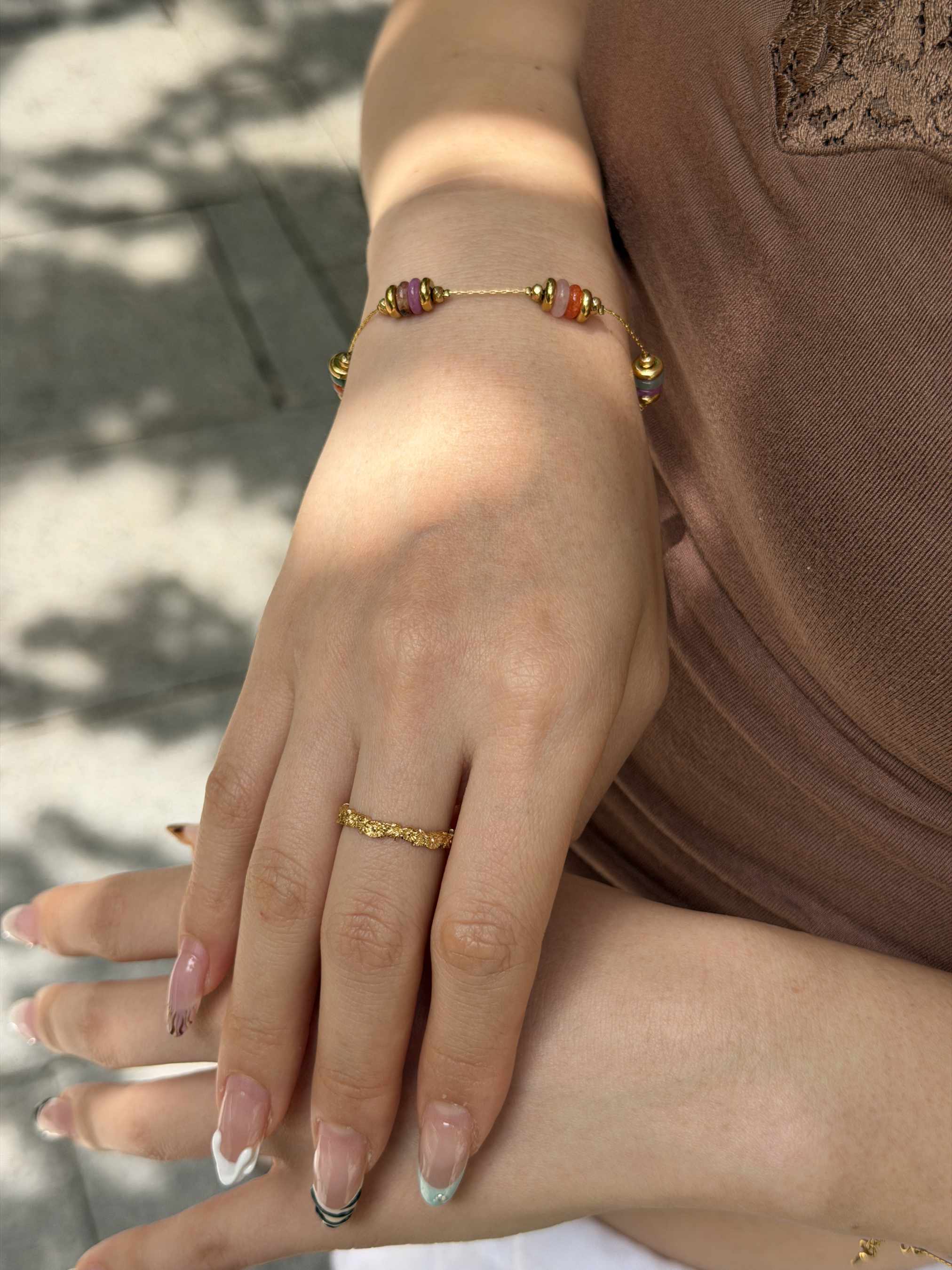 A close-up of a person's hands wearing a delicate gold beaded bracelet on one wrist and a thin gold ring on one finger, set against a brown top and a blurred outdoor background.