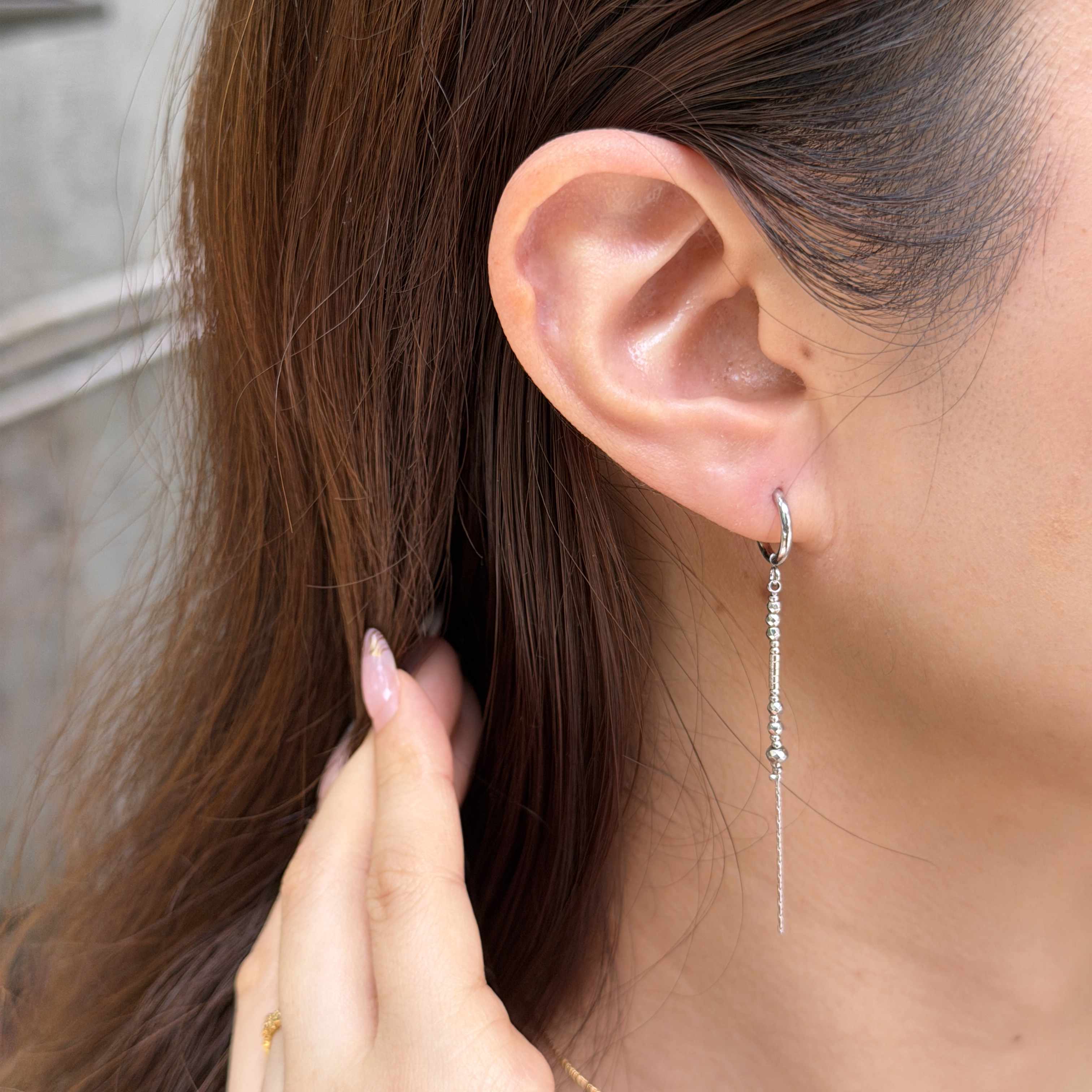 A close-up of a woman's ear wearing a silver huggie hoop earring with a long dangling chain and small clear stones.