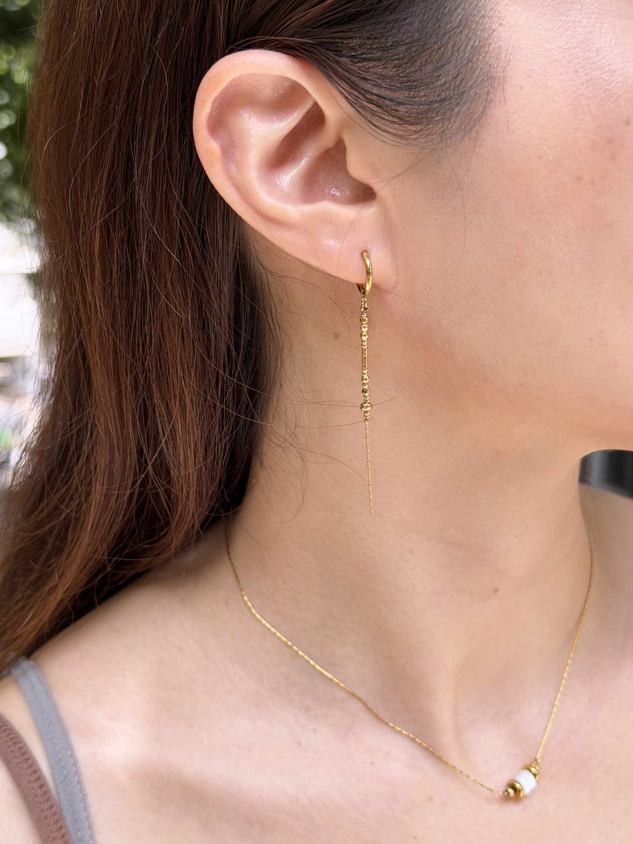 A close-up of a woman's ear wearing a delicate gold drop hoop earring, with a matching thin gold chain necklace also visible.