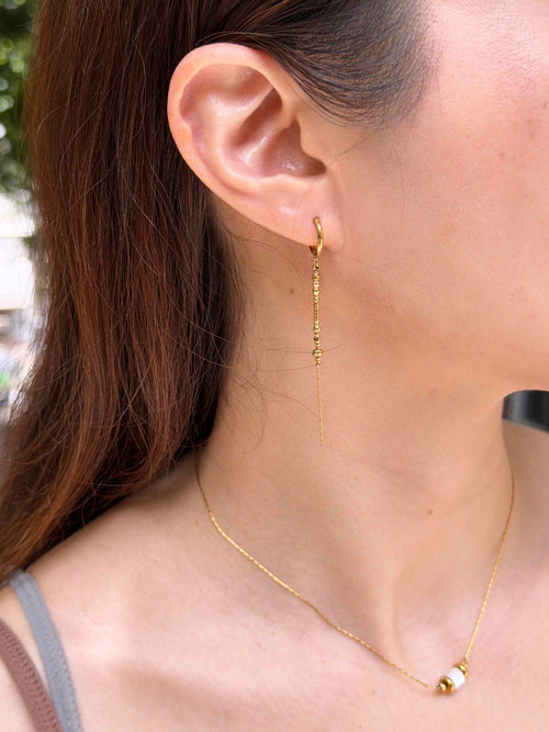 A close-up of a woman's ear wearing a delicate gold drop hoop earring, with a matching thin gold chain necklace also visible.