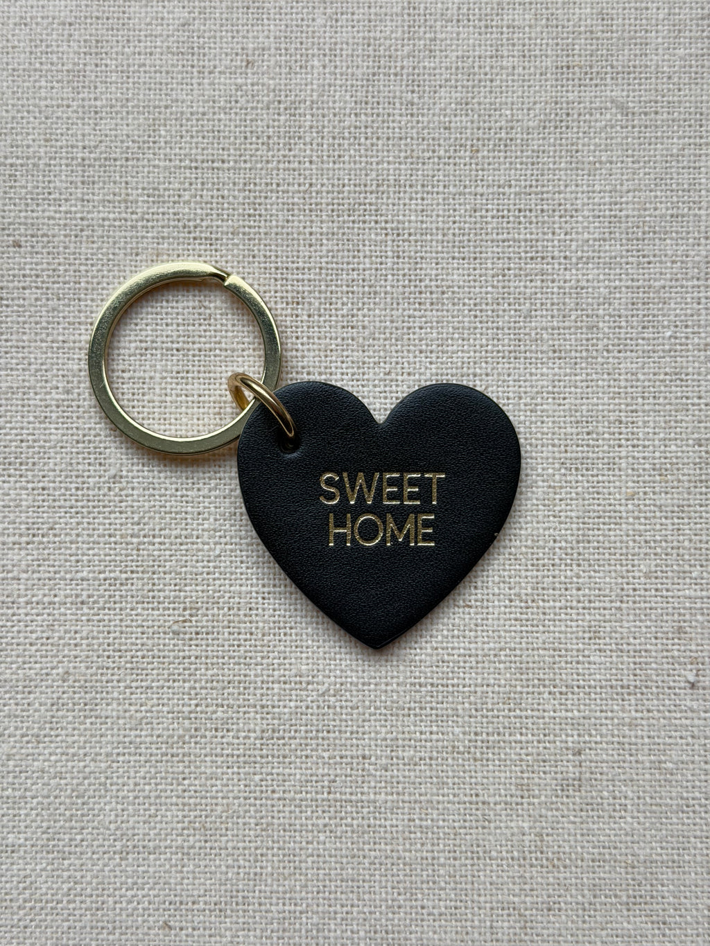Black heart-shaped keychain with 'SWEET HOME' text on a beige fabric background