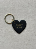 Black heart-shaped keychain with 'SWEET HOME' text on a beige fabric background