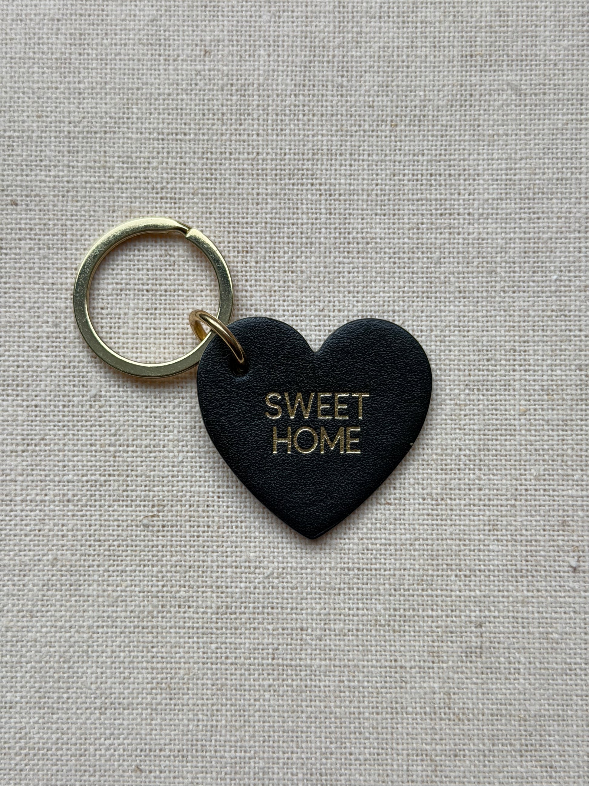Black heart-shaped keychain with 'SWEET HOME' text on a beige fabric background