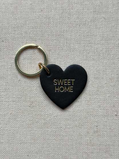 Black heart-shaped keychain with 'SWEET HOME' text on a beige fabric background
