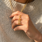 Hand wearing a gold ring with a matching necklace against a neutral background
