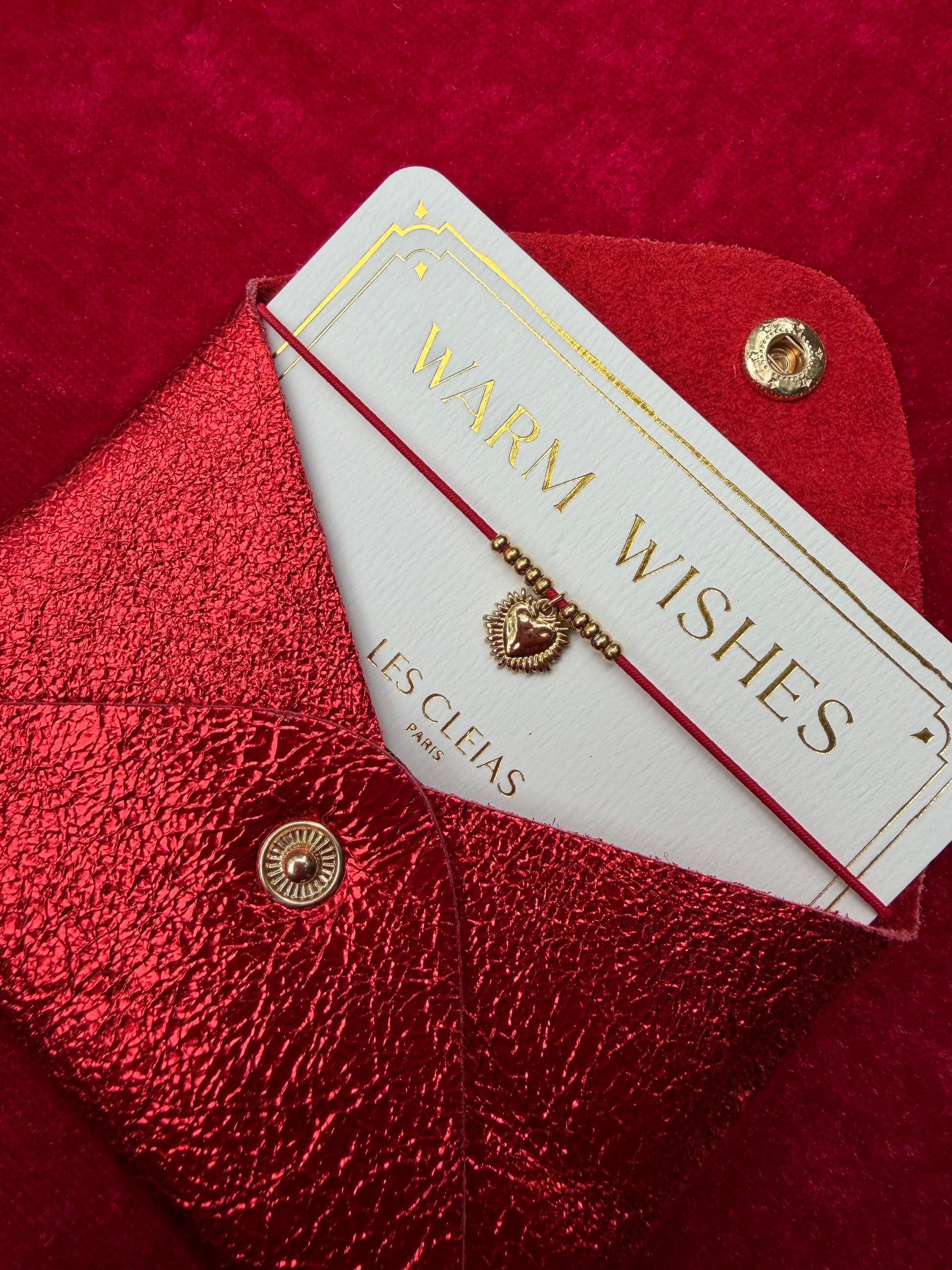 Gold bracelet on a 'Warm Wishes' card with 'LES CLEIAS' branding on a red textured surface.