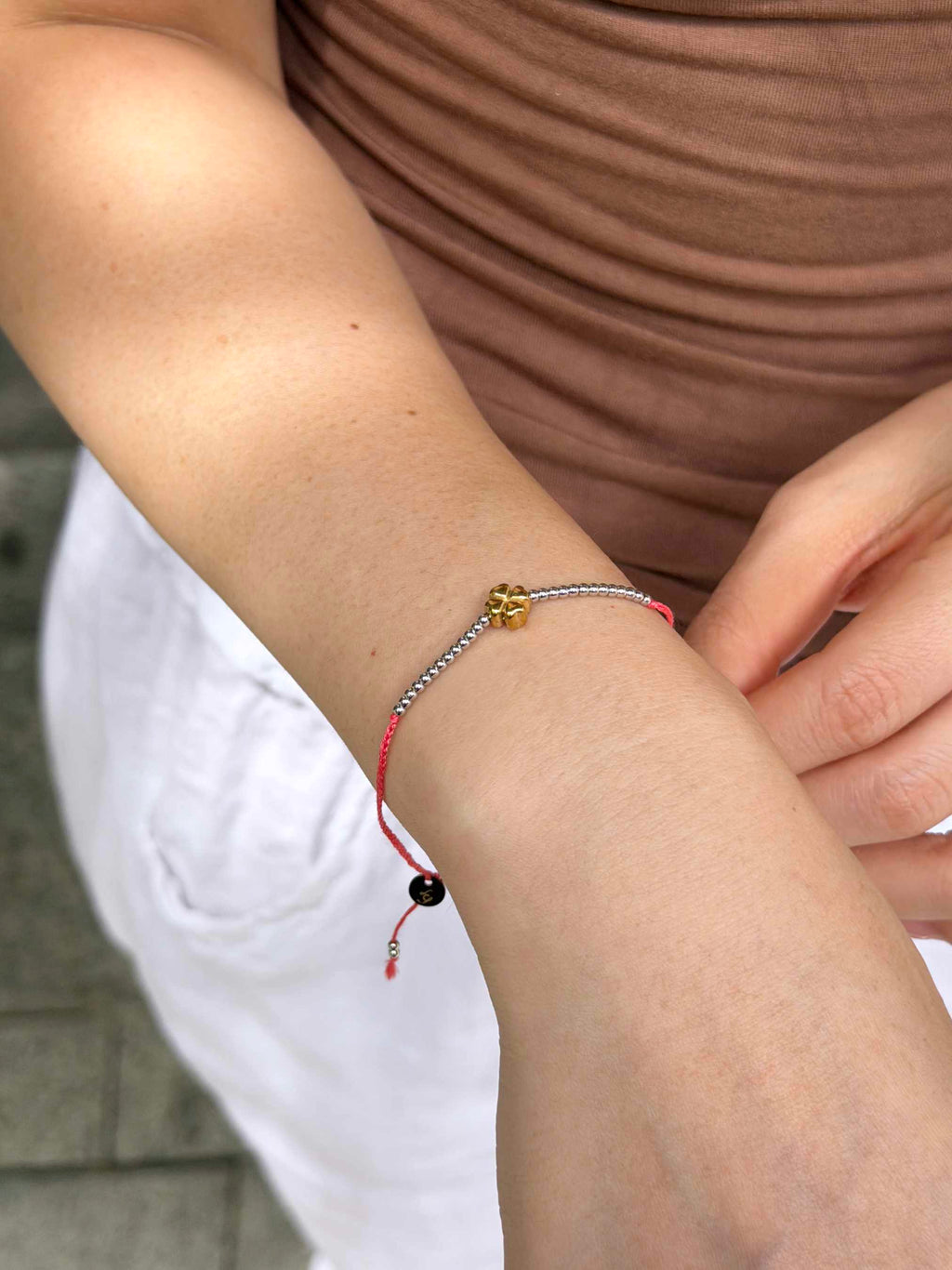 A close-up of a person's wrist wearing a delicate cord bracelet with a small gold charm, a silver-toned beaded section, and a red cord with a black bead.