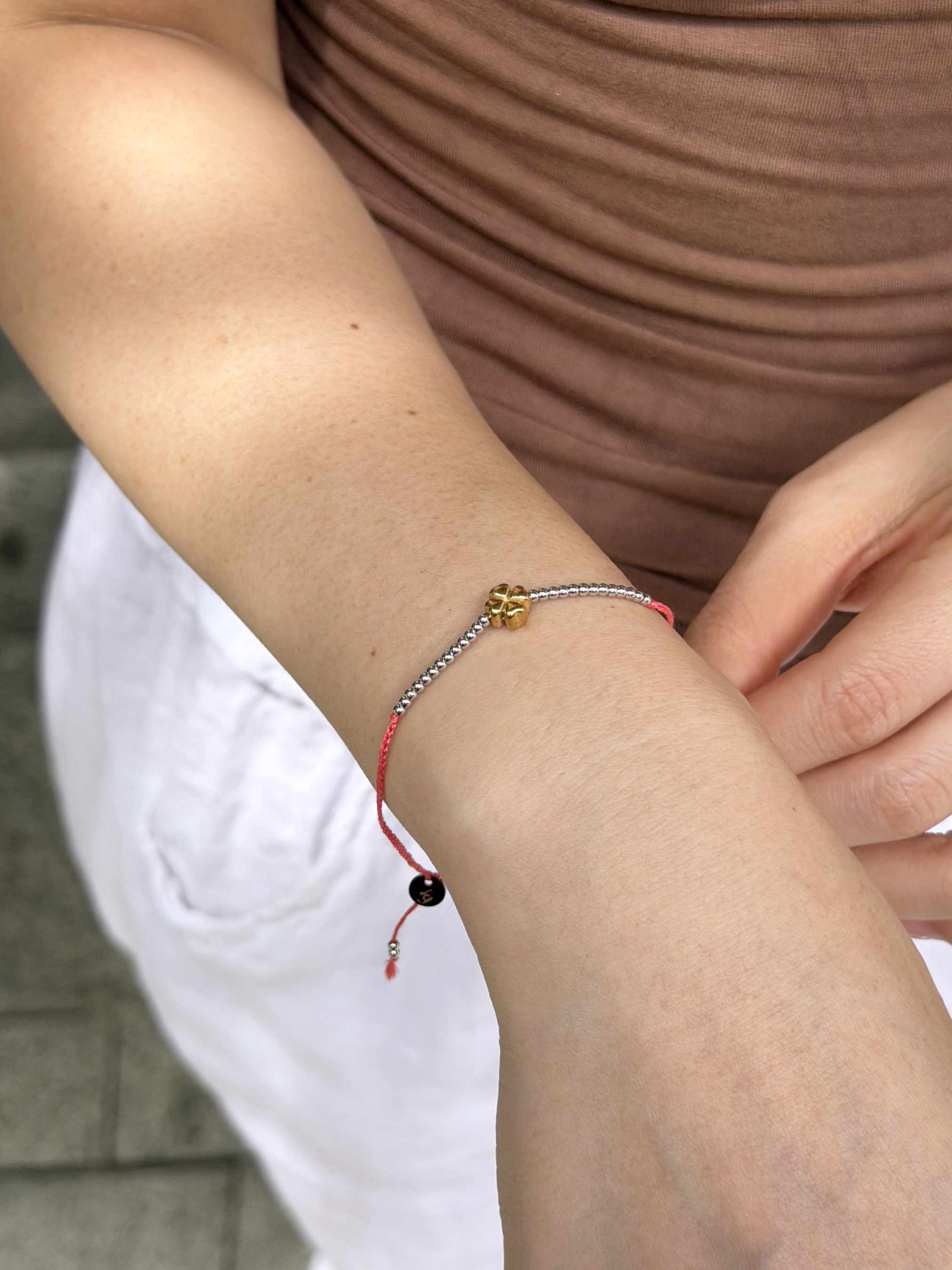 A close-up of a person's wrist wearing a delicate cord bracelet with a small gold charm, a silver-toned beaded section, and a red cord with a black bead.