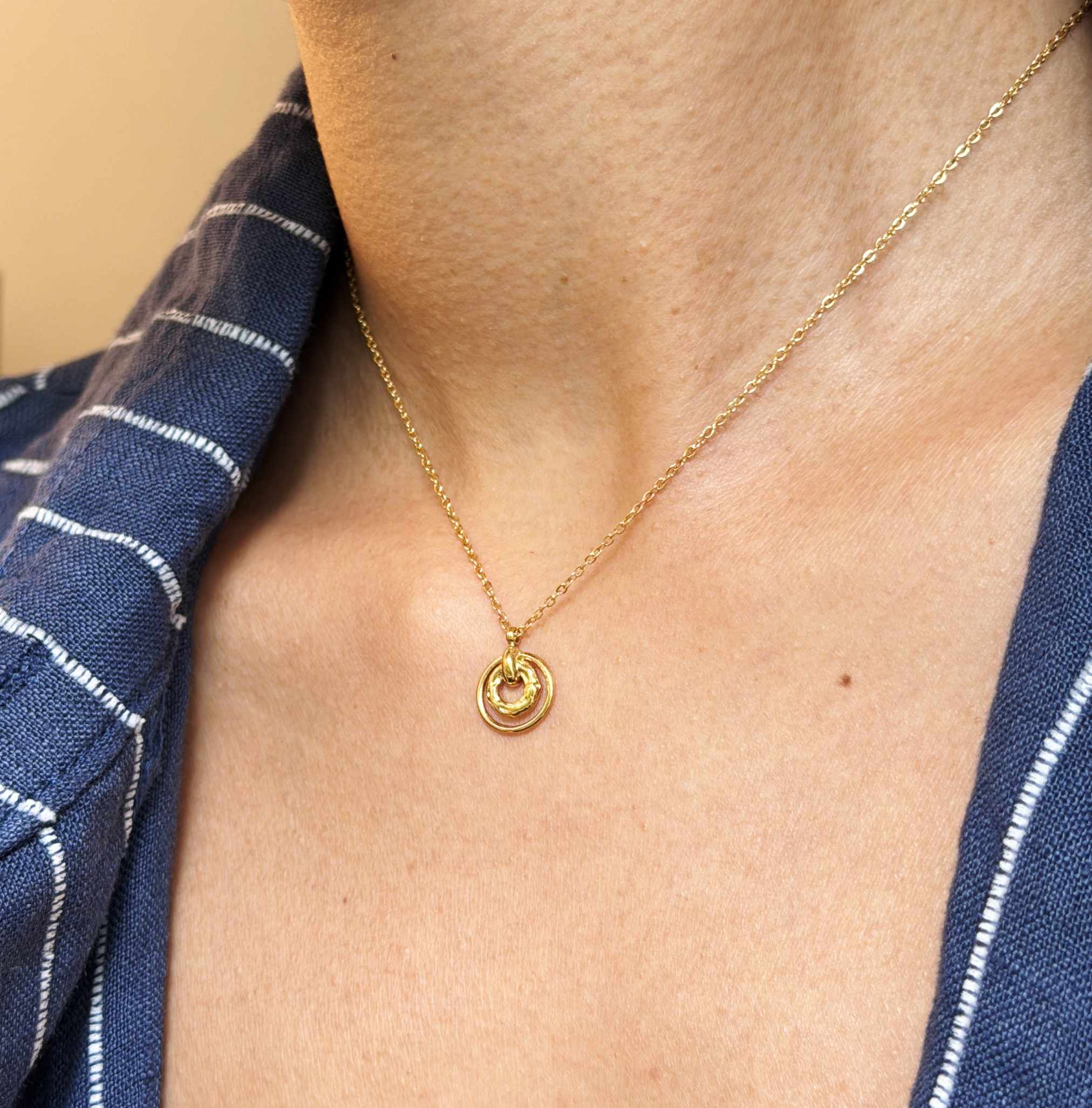 Gold necklace with a circular pendant worn by a person against a blue and white striped fabric background.