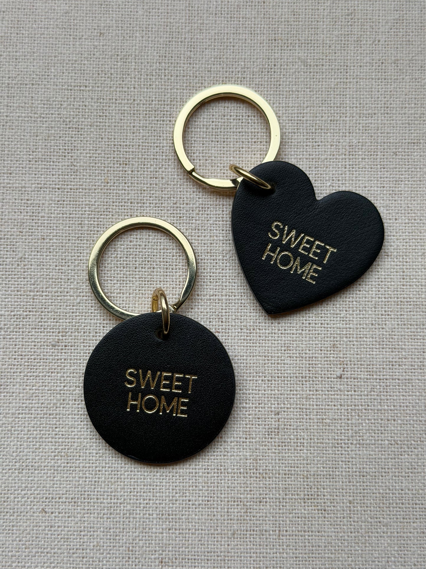 Two black heart-shaped keychains with 'SWEET HOME' text on a beige fabric background