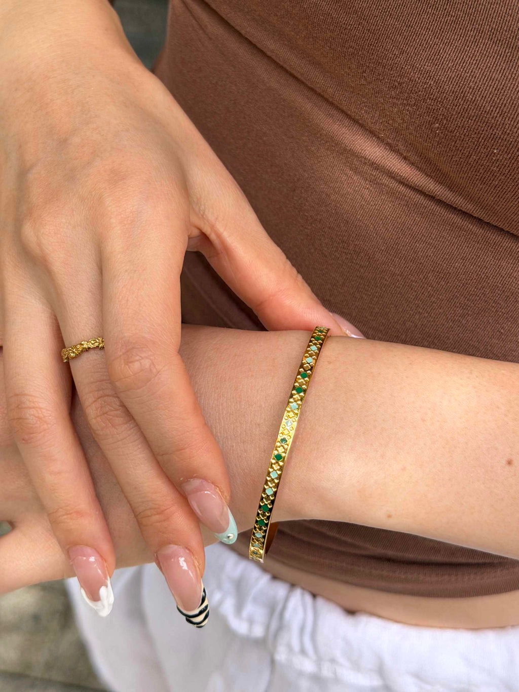 A close-up of a person's wrist wearing a gold bangle bracelet with green and yellow stones, alongside a matching gold ring on their finger.