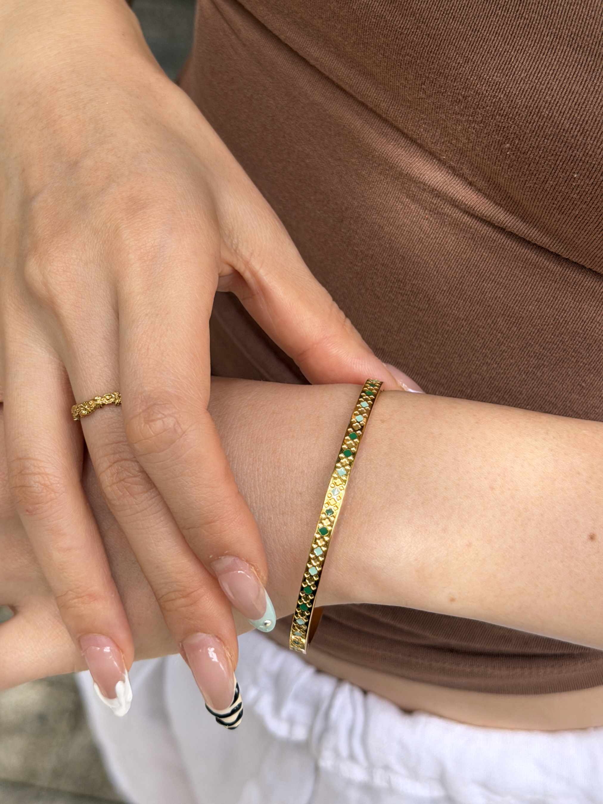 A close-up of a person's wrist wearing a gold bangle bracelet with green and yellow stones, alongside a matching gold ring on their finger.