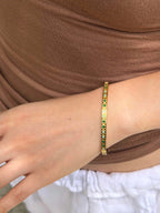 A close-up of a person's wrist wearing a thin gold bangle bracelet with small green and white stones set along the band, paired with a brown top and white bottom.