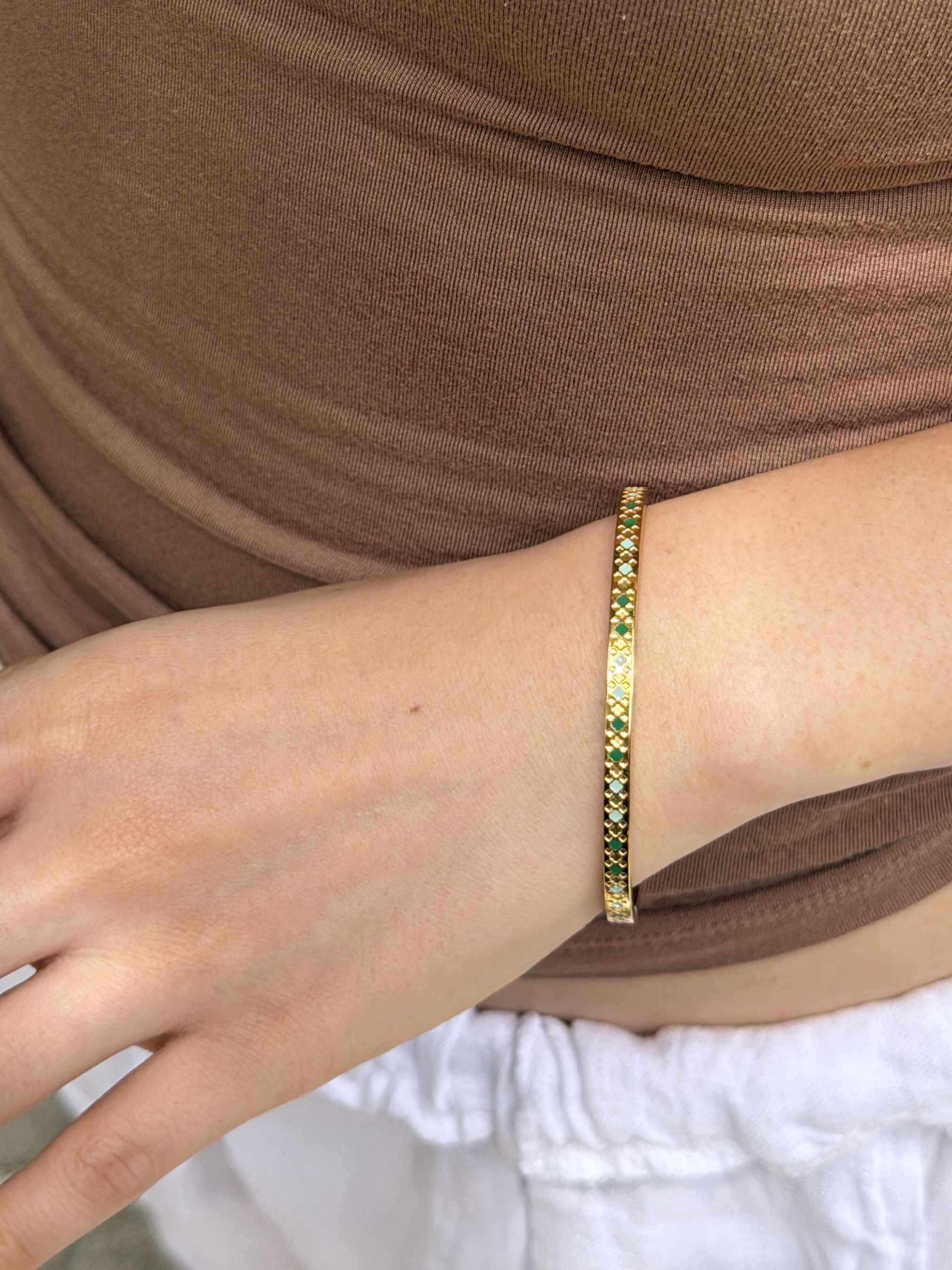 A close-up of a person's wrist wearing a thin gold bangle bracelet with small green and white stones set along the band, paired with a brown top and white bottom.