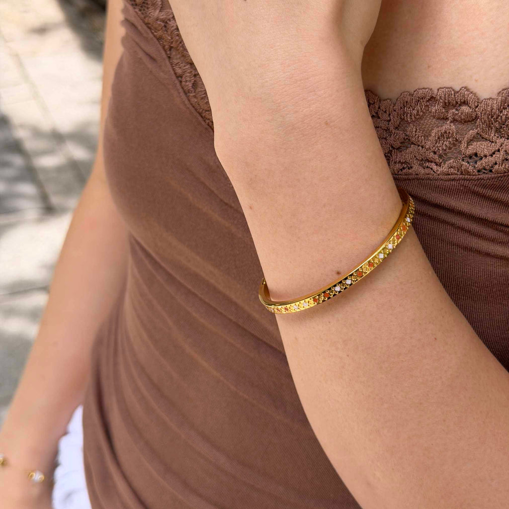 A close-up of a woman's wrist wearing a thin gold bangle bracelet with small clear stones, paired with a brown lace-trim top.