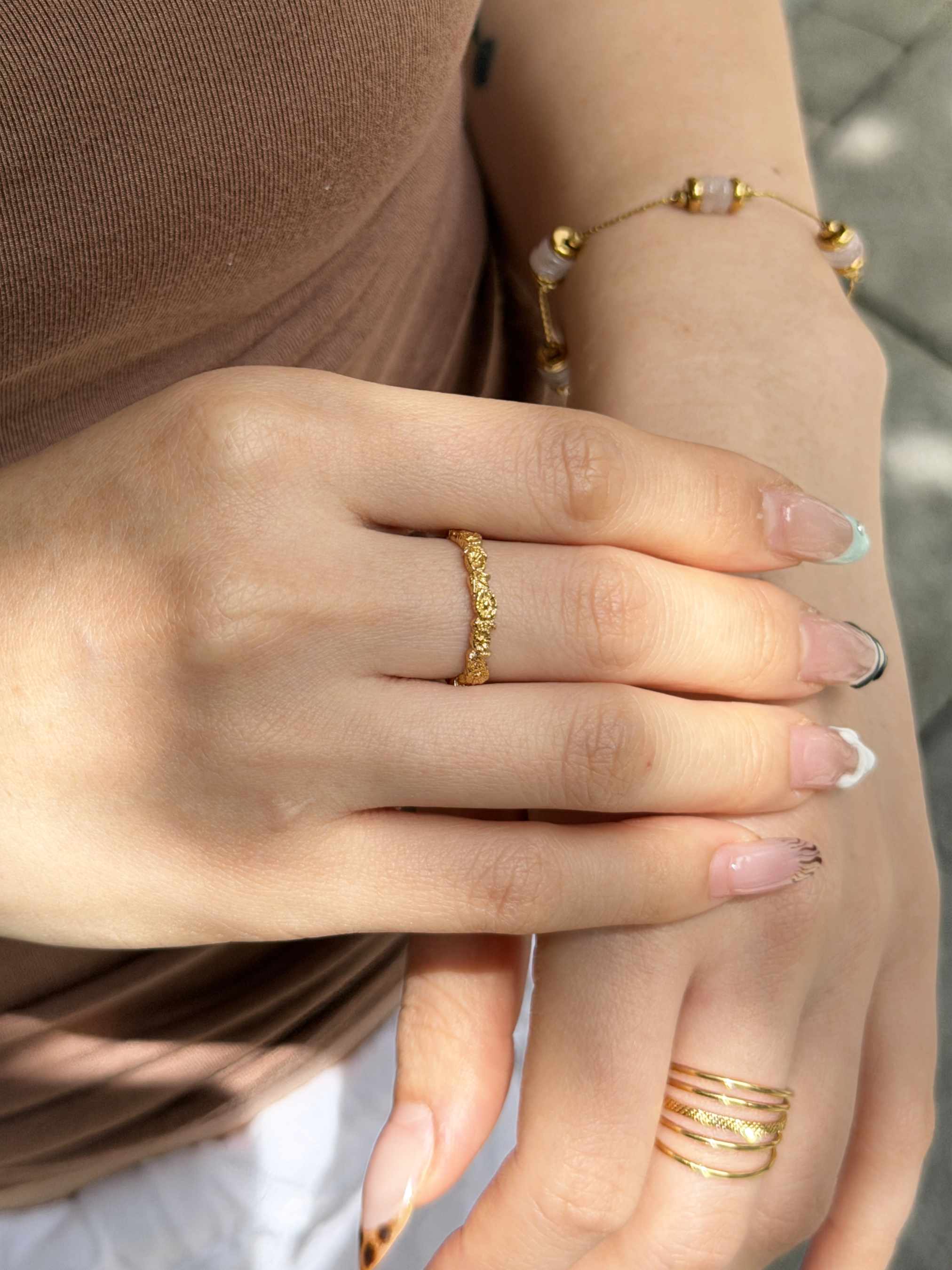Close-up of a person's hands wearing gold jewelry, including a textured gold ring on one finger, a beaded gold bracelet on the wrist, and stacked gold rings on the other hand.